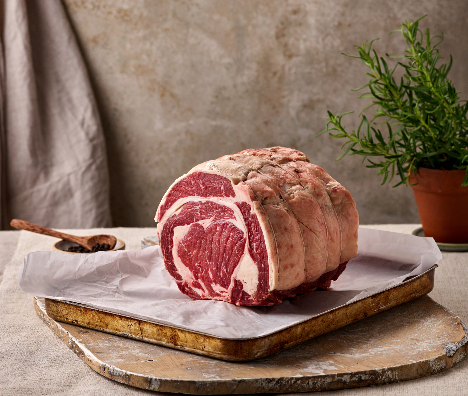 Raw rib roast of heritage breed grass fed beef on a wooden cutting board with a plant in the background, showcasing the ability of our butchers and quality of the beef
